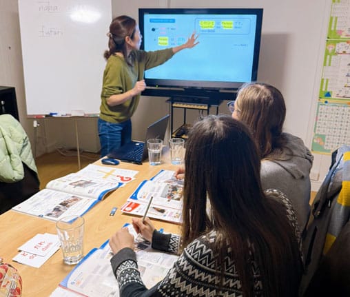 Japanisch Lehrerin zeigt auf der elektronischen Tafel einen Text, zwei Schülerinnen sitzen am Tisch davor