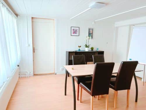 Weißer Raum mit Tisch und vier Stühlen. Im Hintergund ein Sideboard mit ein paar Blumen